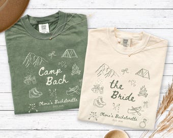 May include: Two crew neck t-shirts, one olive green and one cream, featuring white and dark green line art of camping gear, mountains, and hearts. The text reads Camp Bach and the Bride, with Olivia's Bachelorette July 2026 printed below.