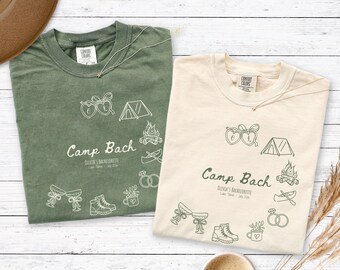 May include: Two crew neck t-shirts in olive green and cream, each featuring a line art graphic with the text "Camp Bach," "Olivia's Bachelorette," and "Lake Tahoe - July 2024," surrounded by illustrations of a tent, campfire, canoe, rings, and boots.