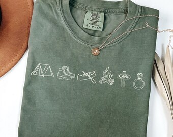 May include: An olive green short-sleeve t-shirt featuring a white line art graphic of a tent, hiking boots, a canoe with an oar, a campfire, a signpost labeled CAMP, and a diamond ring. The shirt has a Comfort Colors brand neck label.