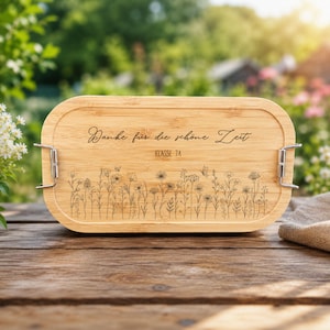Könnte beinhalten: Eine ovale Lunchbox aus Bambus mit heller Holzmaserung. Der Deckel ist mit einem lasergravierten Wiesenblumen-Motiv sowie dem Text "Danke für die schöne Zeit" und "KLASSE 7A" versehen. Zwei seitliche Verschlüsse aus silbernem Metall sichern den Deckel.