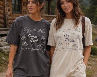 May include: Two short-sleeved t-shirts, one charcoal gray and one beige, featuring a white graphic print of mountains, a tent, a campfire, hiking boots, a canoe, and a ring. The text reads "Camp Bach" or "the Bride" with "Olivia's Bachelorette July 2026".