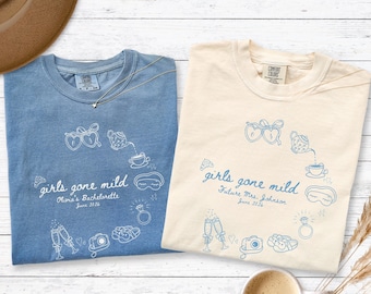 May include: Two crew neck t-shirts in blue and cream, each featuring a blue line art graphic with the text "girls gone mild" and personalized event details. The designs include illustrations of hearts, a teapot, a ring, slippers, and champagne flutes.