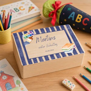 Könnte beinhalten: Eine rechteckige Holzbox mit blau-weiß gestreiftem Rand. In der Mitte steht der Text 'Martins erster Schultag 03.08.2026', ergänzt durch Aquarellillustrationen eines Malkastens, eines Pinsels und eines Stapels bunter Bücher.