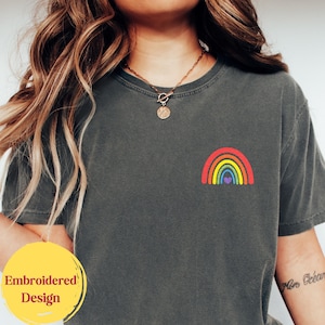 May include: A charcoal gray short-sleeve t-shirt featuring a small, embroidered rainbow graphic on the left chest. The rainbow consists of red, orange, yellow, green, blue, and purple bands with a small purple heart centered at the base.