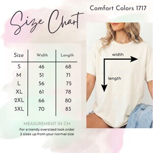 K&ouml;nnte beinhalten: Eine Gr&ouml;&szlig;entabelle f&uuml;r Comfort Colors 1717 T-Shirts mit Ma&szlig;en in Zentimetern. Die Tabelle enth&auml;lt die Gr&ouml;&szlig;en S bis 3XL mit den entsprechenden Breiten- und L&auml;ngenma&szlig;en. Eine Person tr&auml;gt ein cremefarbenes T-Shirt. Der Text auf dem Bild lautet "Size Chart".