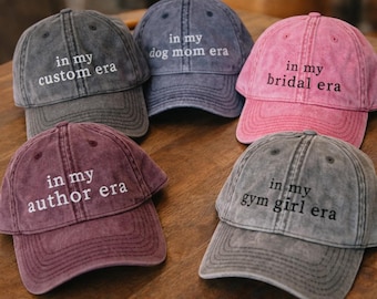 May include: Five washed-out baseball caps in various colors: gray, navy, pink, burgundy, and another gray. Each cap has the phrase "in my" followed by a different era embroidered in white, such as "custom era" or "dog mom era."