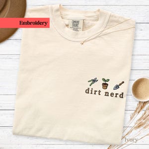 Könnte beinhalten: Elfenbeinfarbenes T-Shirt mit dem Wort "Embroidery" auf einem roten Banner. Das Shirt hat ein gesticktes Design von Gartengeräten und einem Blumentopf mit den Worten "dirt nerd" darunter. Eine Tasse Kaffee und ein Hut sind im Hintergrund.