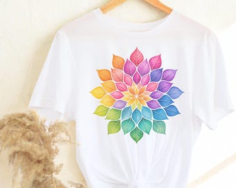 May include: White t-shirt featuring a colorful floral design. The design is a vibrant array of rainbow colors, including orange, yellow, green, blue, and purple, arranged in a leaf-like pattern. The shirt is hanging on a wooden hanger.