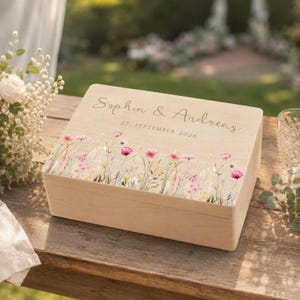 Könnte beinhalten: Eine rechteckige Aufbewahrungsbox aus hellem Holz mit einem aufgedruckten Motiv aus bunten Wildblumen am unteren Rand. Der Deckel ist mit den Namen Sophia & Andreas sowie dem Datum 27. SEPTEMBER 2026 in einer Schreibschrift graviert.