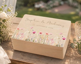 May include: A rectangular light-colored wooden keepsake box featuring a printed design of colorful wildflowers along the bottom edge. The lid is engraved with the names Sophia & Andreas and the date 27. SEPTEMBER 2026 in a script font.