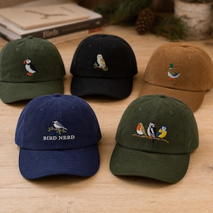 Könnte beinhalten: Fünf Baseballkappen aus Cord in Olivgrün, Schwarz, Hellbraun, Marineblau und Dunkelgrün. Jede Kappe zeigt gestickte Vogelmotive, darunter einen Papageientaucher, eine Eule, eine Stockente und Singvögel. Die marineblaue Kappe trägt den Text BIRD NERD.