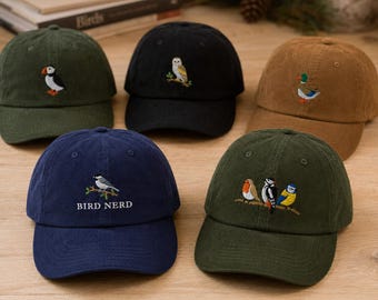May include: Five corduroy baseball caps in olive green, black, tan, navy blue, and dark green. Each cap features embroidered bird designs, including a puffin, owl, mallard, and songbirds. The navy blue cap includes the text BIRD NERD.