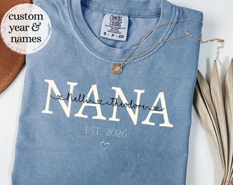 May include: A blue crewneck t-shirt featuring the white text NANA in a large serif font, with the names Nellie and Theodore in black script, the text EST. 2026, and a small heart icon. A gold chain necklace rests on the collar of the shirt.