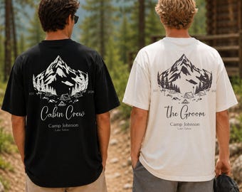 May include: A black t-shirt and a white t-shirt, each featuring a back graphic of a mountain range, pine trees, and tents. The black shirt displays the text Cabin Crew, Camp Johnson, and Lake Tahoe. The white shirt displays The Groom, Camp Johnson, and Lake Tahoe.