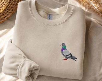 May include: A beige crewneck sweatshirt featuring a small, detailed embroidered pigeon on the left chest. The bird design displays realistic gray, green, and purple plumage with bright red feet. The garment has a ribbed collar and cuffs.