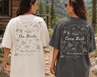 May include: Two oversized t-shirts, one cream and one charcoal gray, featuring a back graphic print with line art of mountains, a tent, a canoe, a campfire, and a ring. The cream shirt says the Bride, and the gray shirt says Camp Bach.