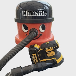 Pode incluir: Um aspirador Numatic vermelho e preto com um rosto sorridente, uma mangueira preta e uma lixadeira DeWalt preta e amarela com uma bateria. O aspirador tem a palavra "Numatic" impressa no topo.
