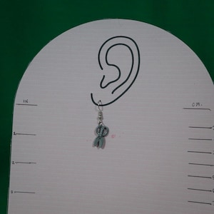 May include: A light blue scissor-shaped earring with a silver hook. The earring is displayed on a white surface with a drawn ear and centimetre measurements.