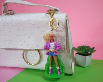 Könnte beinhalten: Eine weiße Handtasche mit goldenen Details und einem Puppen-Schlüsselanhänger. Die Puppe trägt eine rosa Jacke, blaue Shorts und rosa Stiefel. Der Schlüsselanhänger ist an der Tasche befestigt. Rosa Hintergrund.