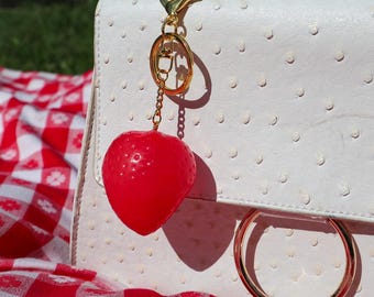 May include: A red heart-shaped keychain charm hangs from a gold-colored clasp attached to a white handbag. The handbag has a textured surface with small, evenly spaced dots. A red and white checkered picnic blanket is in the background.