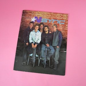 May include: A vintage *NSYNC poster featuring the boy band posing in front of a brick wall. The poster has a blue and white logo with the band's name. The band members are wearing denim jackets, vests, and jeans.