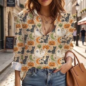 May include: A long-sleeve button-down shirt featuring a repeating Halloween-themed pattern. The print includes black cats, orange jack-o'-lanterns, white ghosts, and green crescent moons on a light beige background with a collared neckline.