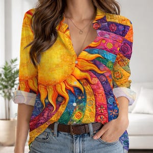 Op de afbeelding: Een button-down shirt met een levendig zon- en regenboogontwerp. Het shirt heeft een grote gele zon aan de linkerkant en een kleurrijk regenboogpatroon aan de rechterkant. De lange mouwen zijn opgerold en het shirt wordt gedragen met een blauwe spijkerbroek en een bruine riem.