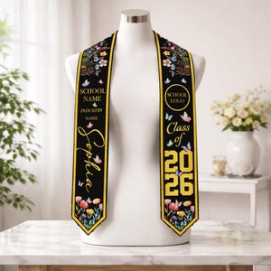 Op de afbeelding: Een zwarte afstudeer sjerp met bloemen- en vlindermotieven en gouden randen. De sjerp toont de naam "Sophia" in gouden letters, evenals de tekst "School Name", "Industry Name", "School Logo" en "Class of 2026".