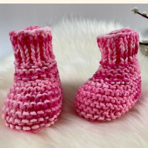 Pode incluir: Um par de botinhas de bebê de tricô rosa e branco. As botinhas são feitas com um ponto aran texturizado e têm uma borda branca ao redor da parte superior. As botinhas estão sobre uma superfície branca e fofa. O texto "PDF Knitting Pattern No Pick Up Newborn Booties in Aran Yarn by Knitting Times Design" é visível na parte superior da imagem.