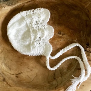May include: White crocheted baby bonnet with a lace trim and a long white crocheted tie. The bonnet is lying on a wooden surface.