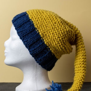 May include: A yellow and blue knitted hat with a long tail. The hat has a blue brim and a yellow top with a blue pom-pom at the end of the tail.