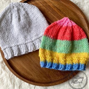 May include: Two knitted beanies. One is plain white, the other is striped with bands of blue, yellow, green, red, and pink. The hats are displayed on a wooden tray. The image has a soft, warm aesthetic.