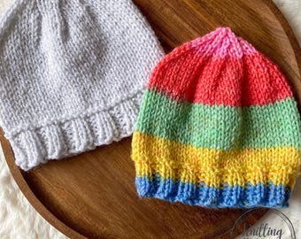 May include: Two knitted beanies. One is solid white, the other is striped with bands of blue, yellow, green, red, and pink. The hats are displayed on a wooden tray. The image has a soft, warm aesthetic.