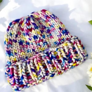 May include: A colorful knitted beanie hat with a chunky knit texture. The hat is made with a variety of colors, including pink, blue, yellow, and white.