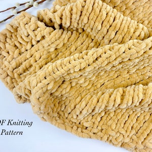 May include: A close-up of a knitted blanket in a light brown colour. The blanket is made with a chunky yarn and has a soft, fluffy texture. The text "PDF Knitting Pattern" is visible in the bottom left corner of the image.