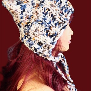 May include: A chunky knit hat in a mix of white, blue, and brown yarn. The hat has a pointed top and braided ties. The hat is shown on a mannequin with red hair, against a dark red background. The hat is a winter accessory.