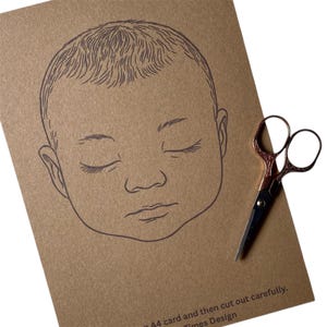 May include: A brown card with a line drawing of a sleeping baby's face. The image is accompanied by a pair of small, decorative scissors with a copper-colored handle. Text at the bottom reads "A4 card and then cut out carefully. Times Design."