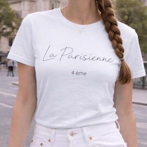 Może przedstawiać: Biała koszulka z napisem "La Parisienne" i "4 ème" w czarnym skrypcie. Koszulka jest połączona z białymi dżinsami. Tło przedstawia ulicę miasta. Zdjęcie zrobiono na zewnątrz.