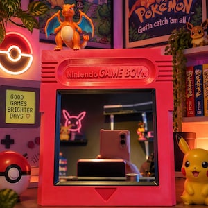 Può includere: Uno specchio rettangolare rosa a forma di cartuccia di gioco Nintendo Game Boy. La cornice presenta il testo in rilievo Nintendo GAME BOY nella parte superiore e un intaglio triangolare nella parte inferiore. La superficie è riflettente.