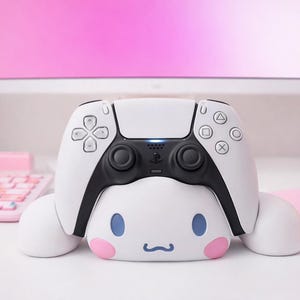 Può includere: Un controller PlayStation 5 bianco e nero poggia su un supporto bianco a forma di personaggio dei cartoni animati con occhi blu e guance rosa. Il controller è su una scrivania bianca con tastiera, mouse e monitor rosa sullo sfondo.