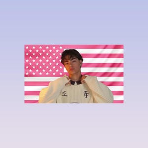 May include: A person wearing a cream-coloured sweater with the letters "FF" on the front, posing in front of a pink and white American flag. The sweater has a ribbed texture and a contrasting collar.