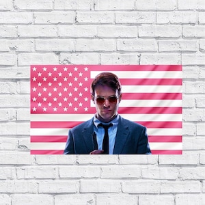 May include: A poster featuring a man in a suit and sunglasses, set against a pink American flag. The man is holding a black object. The background is a white brick wall.