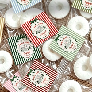 May include: A collection of individually wrapped white ring-shaped candies featuring square labels with colorful vertical stripes. Each label displays a central illustration of a house, the text Camila & Mateo, Engaged, and 12.25.26.