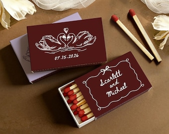 May include: Two rectangular matchboxes with dark maroon covers. One box features a white line drawing of two swans forming a heart shape above the date 07.15.2026. The other box shows the text Scarlett and Michael inside a white decorative frame.