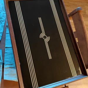 May include: Rectangular serving tray with a dark background and a brown wooden frame. The tray features white vertical stripes and a central geometric design. The tray has two wooden handles and is likely used for serving food or drinks.