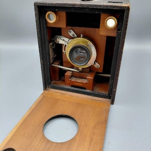 May include: Antique wooden box camera with a brass lens and a hinged front panel. The camera has a leather handle and a dark finish. The lens is centered on the front of the camera, and the front panel has a circular opening.