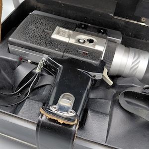 Puede incluir: Cámara de cine Super 8 vintage en negro y plateado, con asa y lente de cuero, dentro de un estuche negro. La cámara tiene una lente plateada y diales de control. El estuche está abierto, revelando un interior negro.
