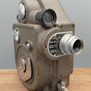 Könnte beinhalten: Vintage braune Filmkamera mit silbernem Objektiv und verschiedenen Einstellrädern. Die Kamera hat ein strukturiertes Gehäuse und ein Etikett mit Belichtungseinstellungen. Ein beiges Kabel erstreckt sich von der Basis. Ein klassisches Fotoequipment.