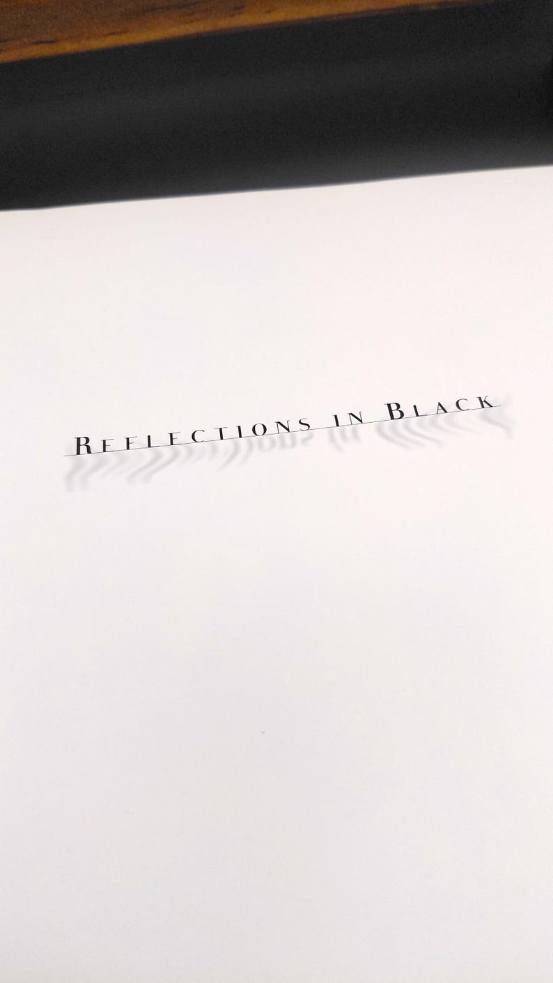 Puede incluir: Superficie blanca con el texto "REFLECTIONS IN BLACK" impreso en una fuente fina y elegante. El texto proyecta una sombra, a&ntilde;adiendo profundidad. Un borde de madera oscura es visible en la parte superior.