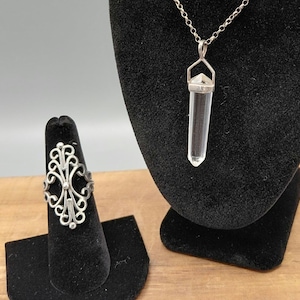 Könnte beinhalten: Ein silberner Filigranring auf einem schwarzen Präsentationsständer. Eine klare Kristall-Anhänger-Halskette hängt an einer schwarzen Samtbüste. Der Ring hat ein verziertes, symmetrisches Design. Der Kristallanhänger hat eine klare, längliche Form mit silbernen Akzenten.